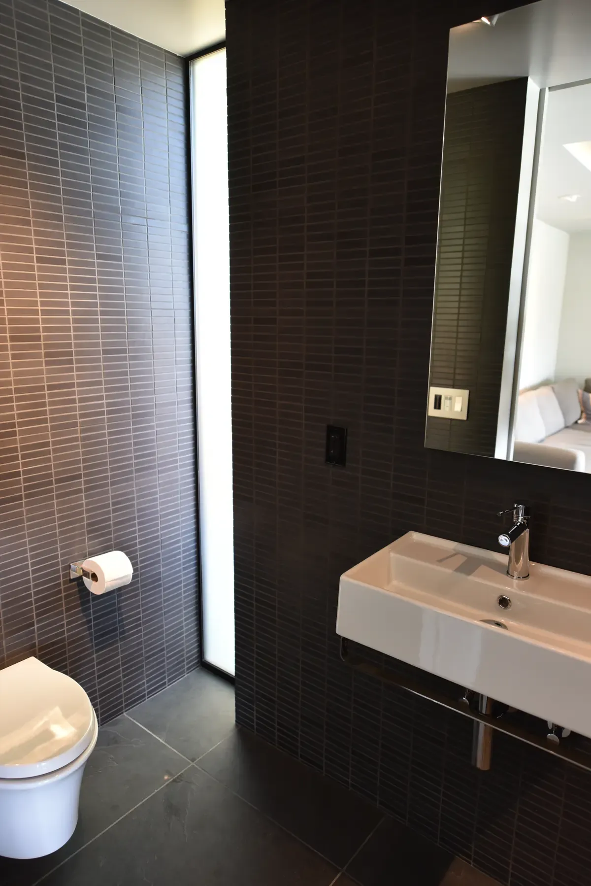 Washroom with a wall-hung basin against dark tile, tall slot window alongside