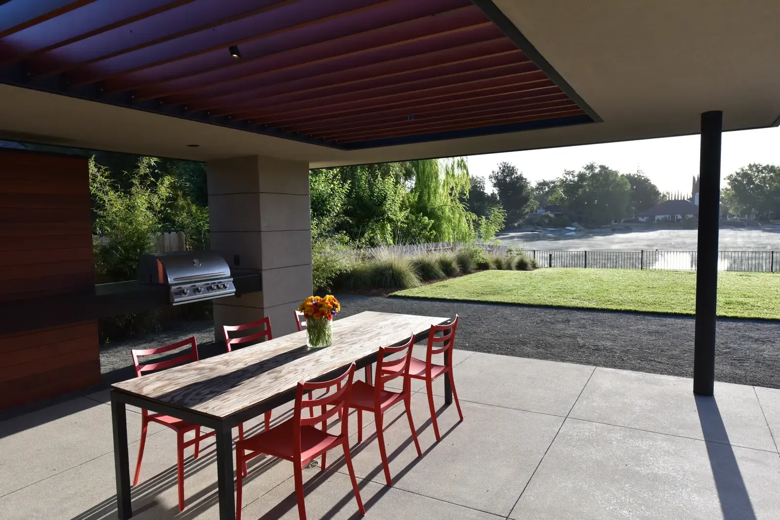 Outdoor kitchen and long dining table with red stools beneath the timber canopy