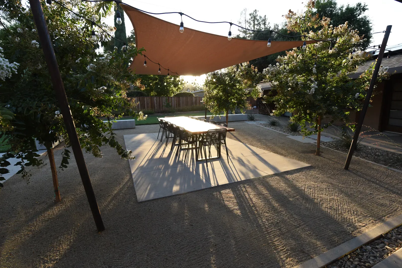 Shade sail and dining table on the gravel terrace, low sun raking across the ground