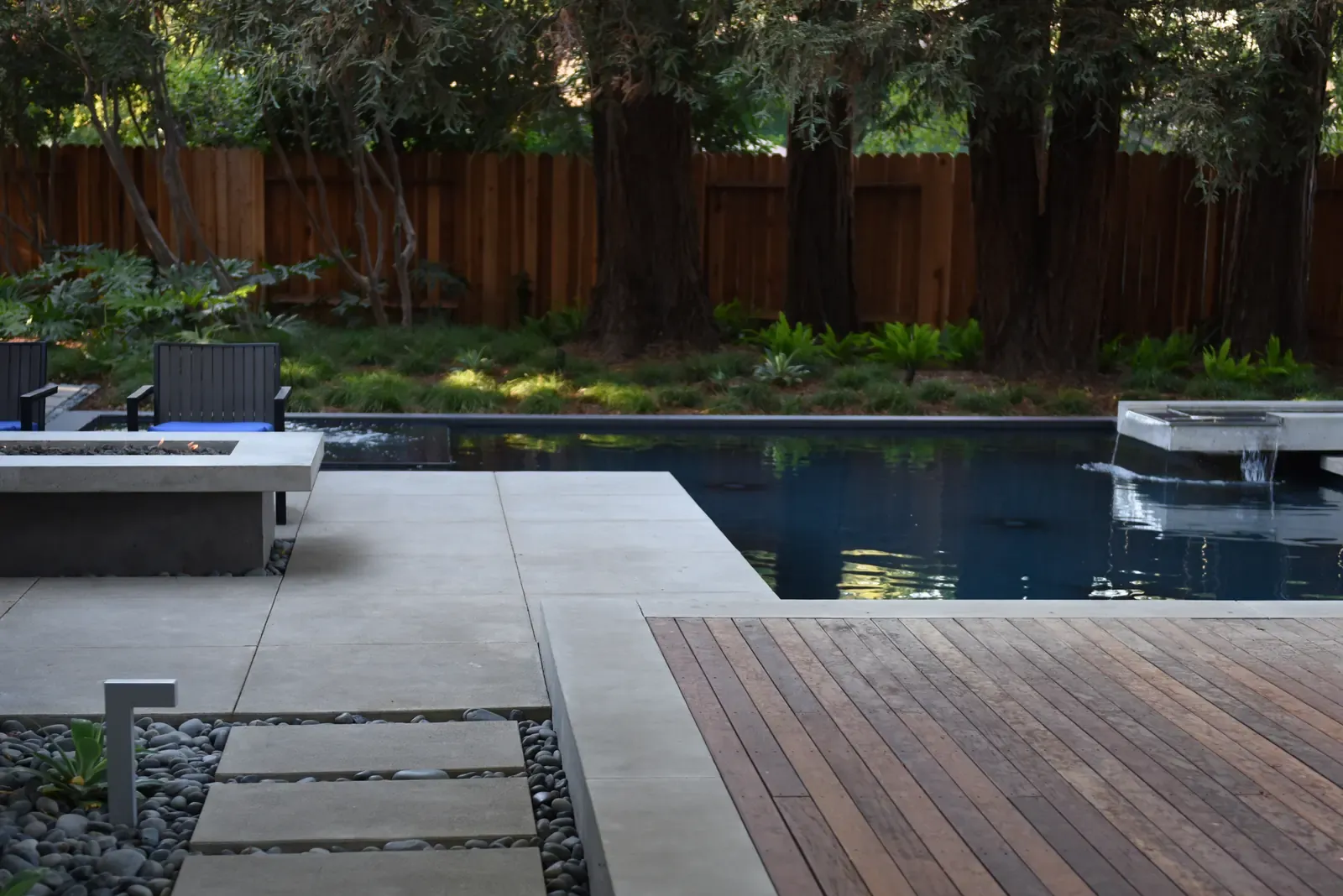 Fire bowl at the pool edge, concrete paving meeting an ipe deck against a redwood fence