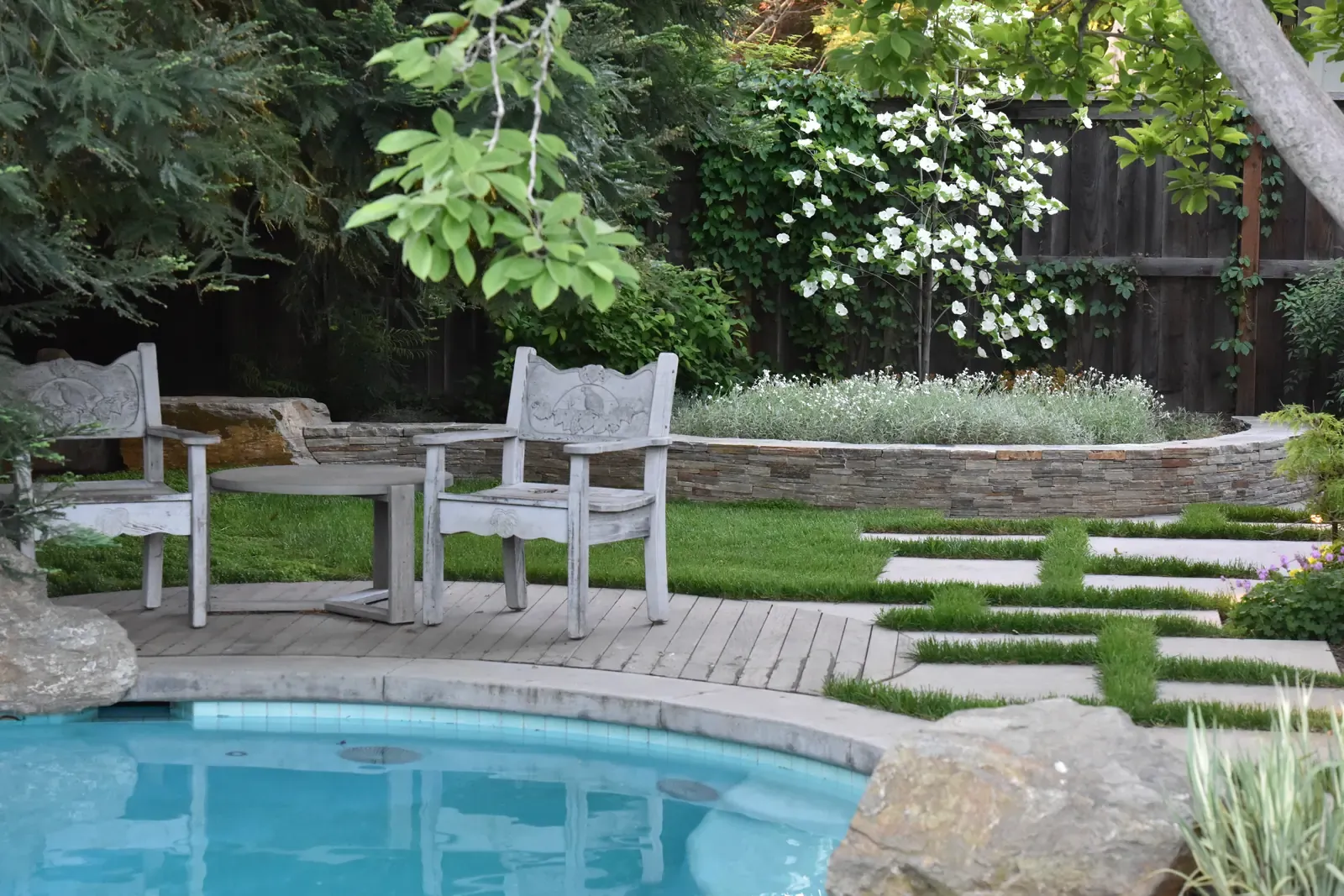 Pair of teak chairs on the lawn beside a raised stone planter, pool edge below