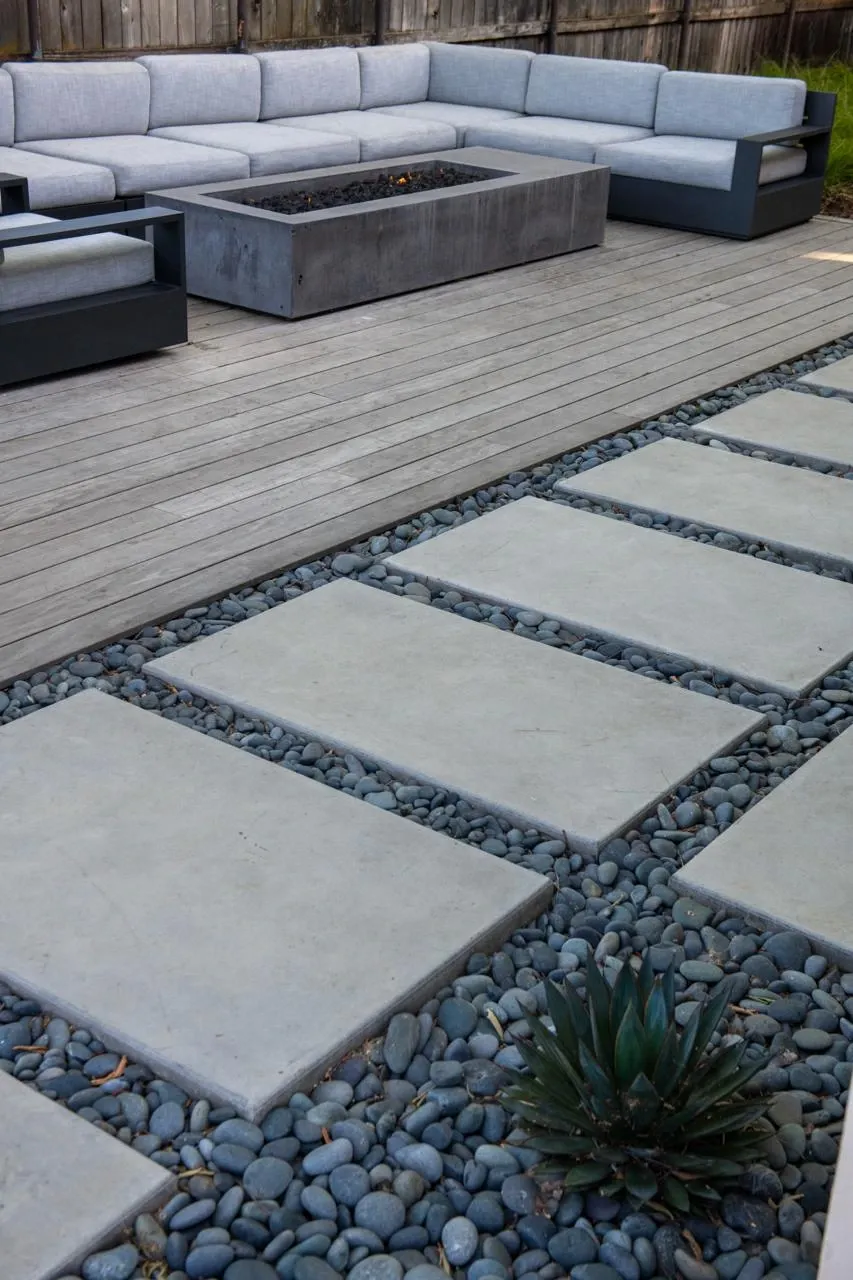 Concrete stepping pads crossing river stones beside a wood deck and fire seating area
