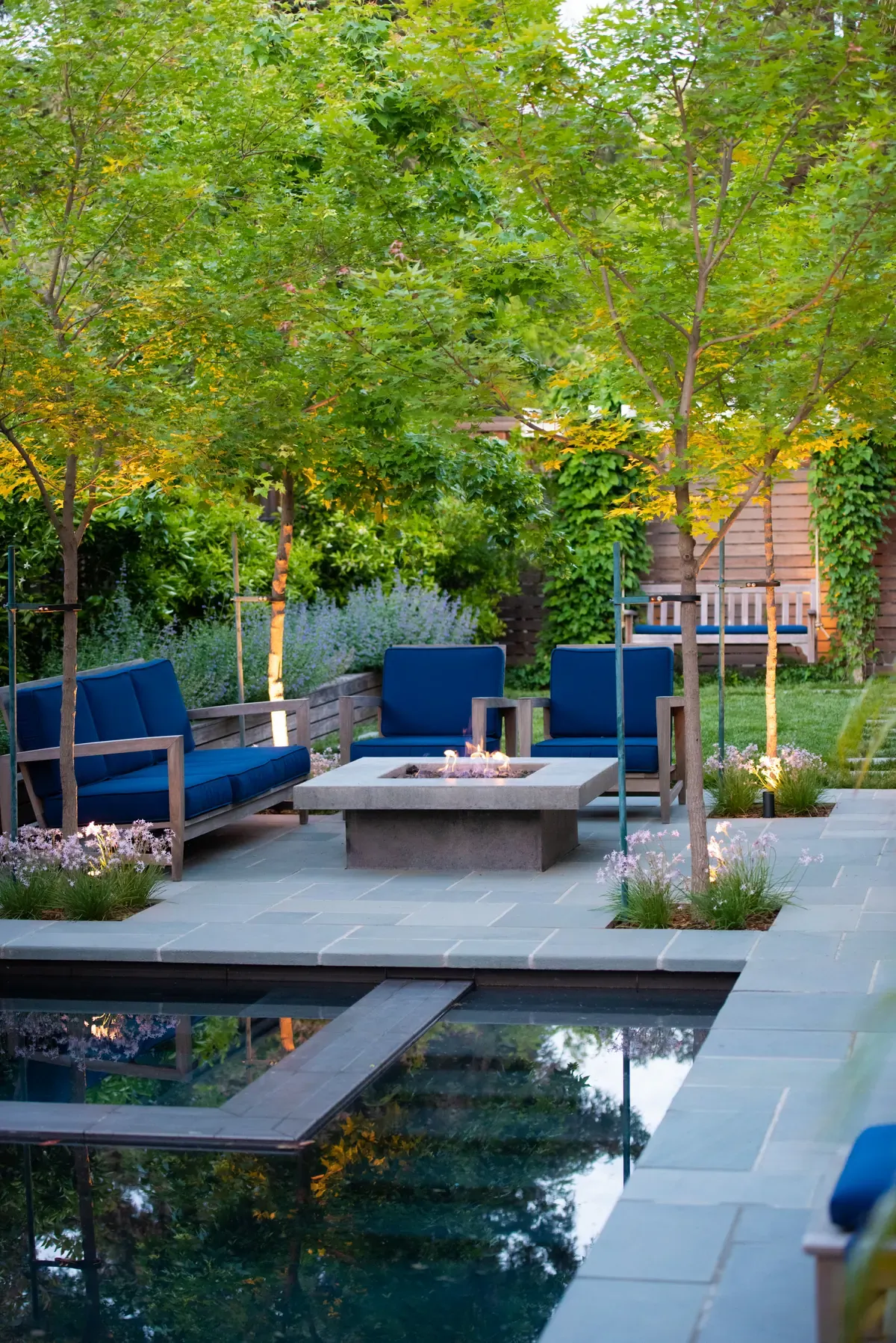 Fire table and blue seating on the terrace, pool reflecting the trees