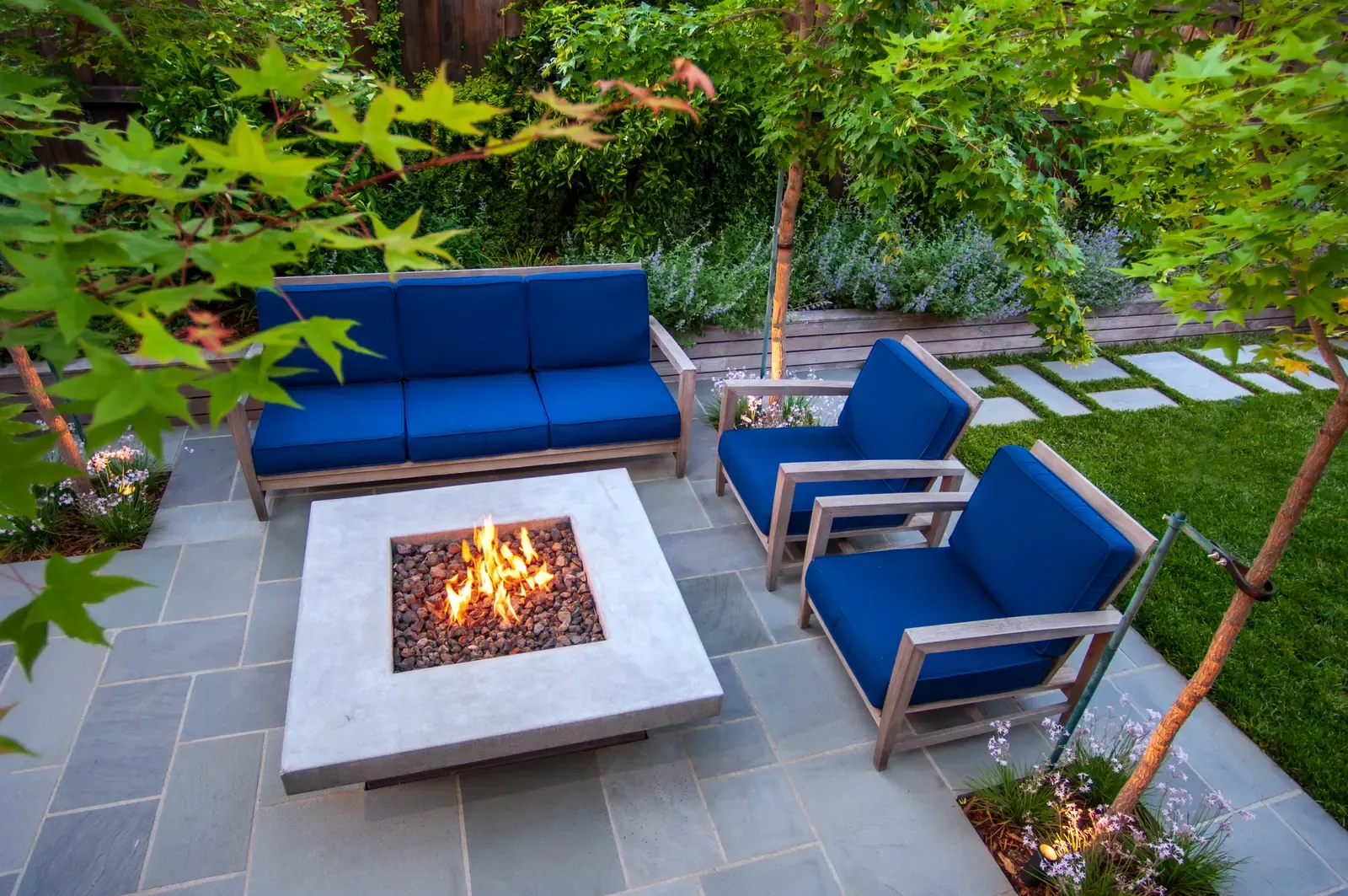Fire table and blue seating under the maple, lawn beyond the paving