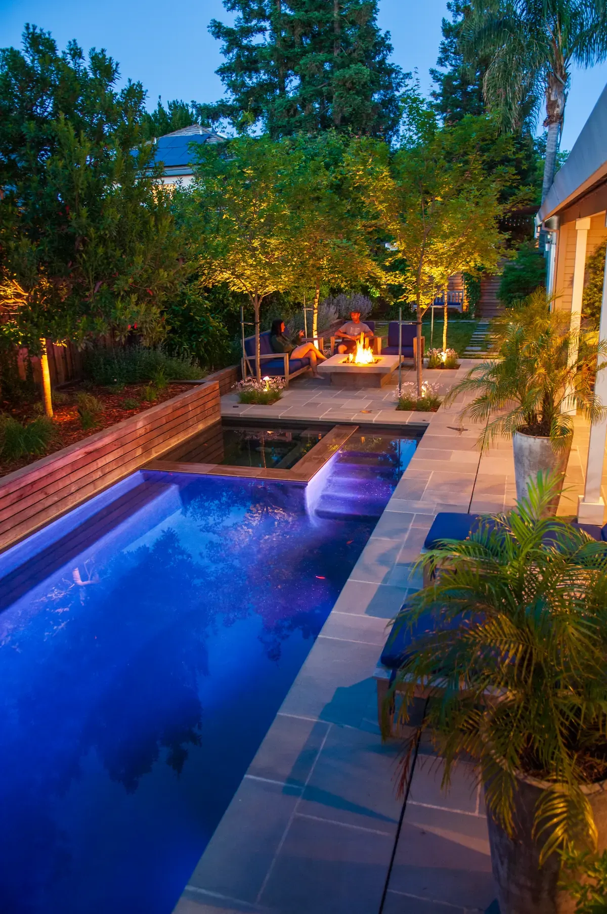 Pool glowing at night, fire and uplit trees beyond