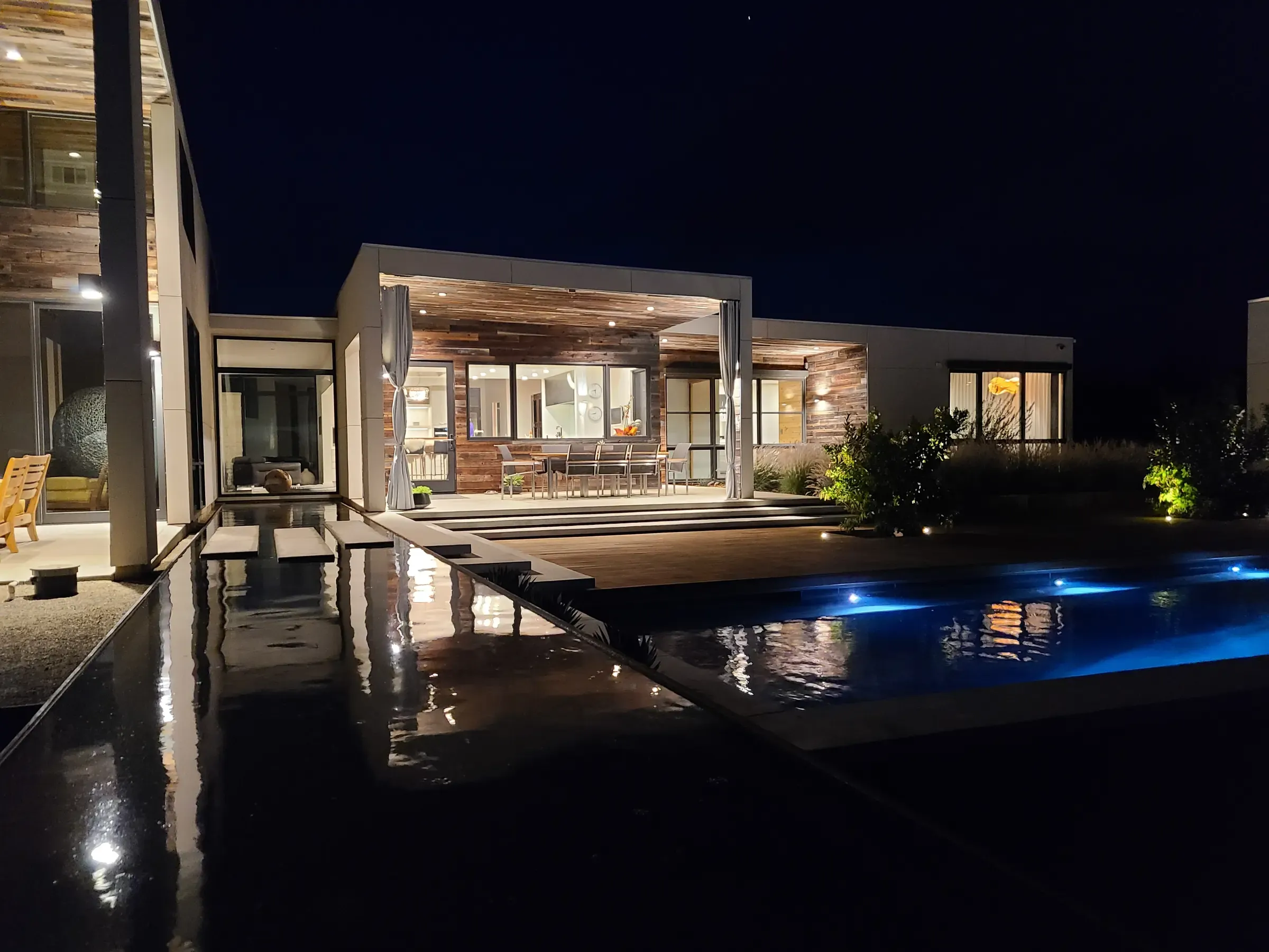 The house lit at night, reflected in the pool with the deck running to the water