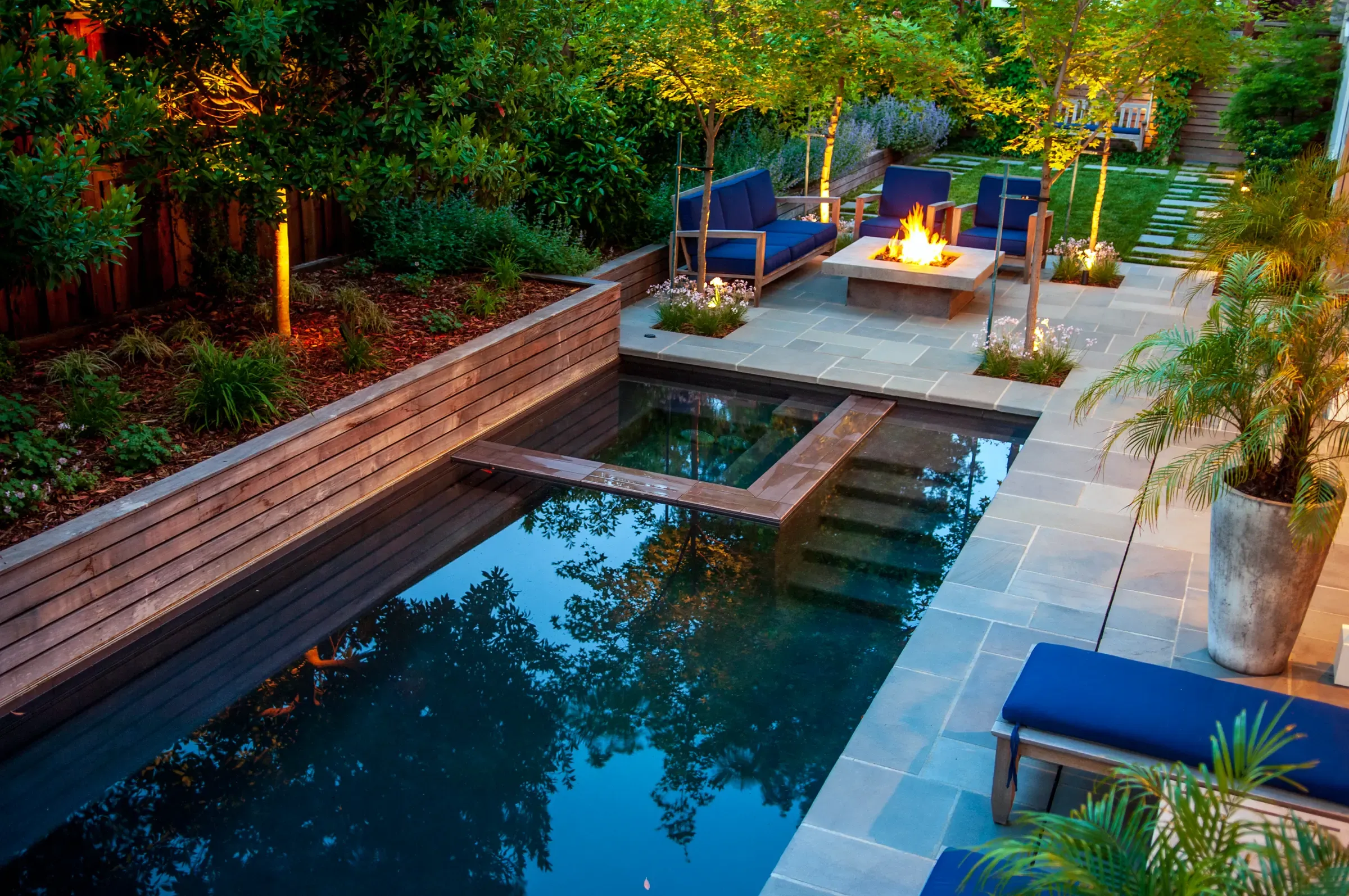 Pool and fire table lit at dusk, blue seating on the bluestone and a timber wall behind