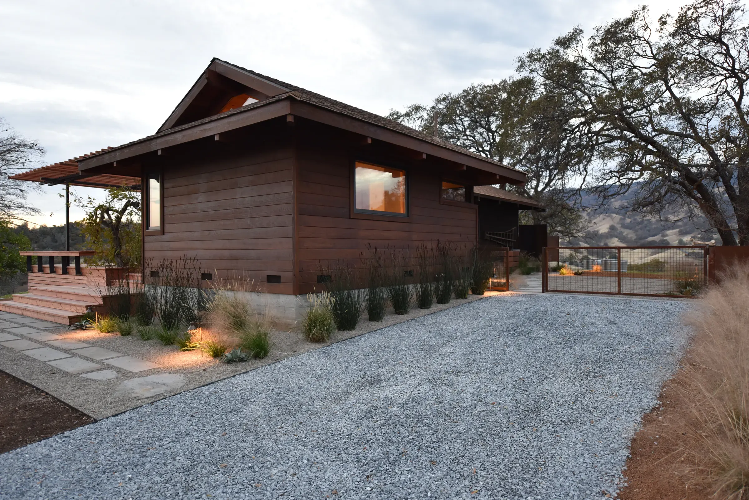 Timber-clad addition at dusk, gravel drive and grasses running to a valley oak